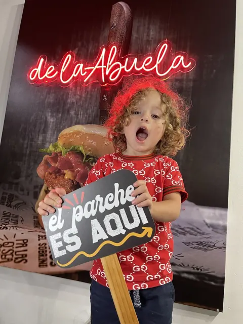 Niño con el cartel «el parche es aquí» frente al letrero de neón de la hamburguesa De la Abuela